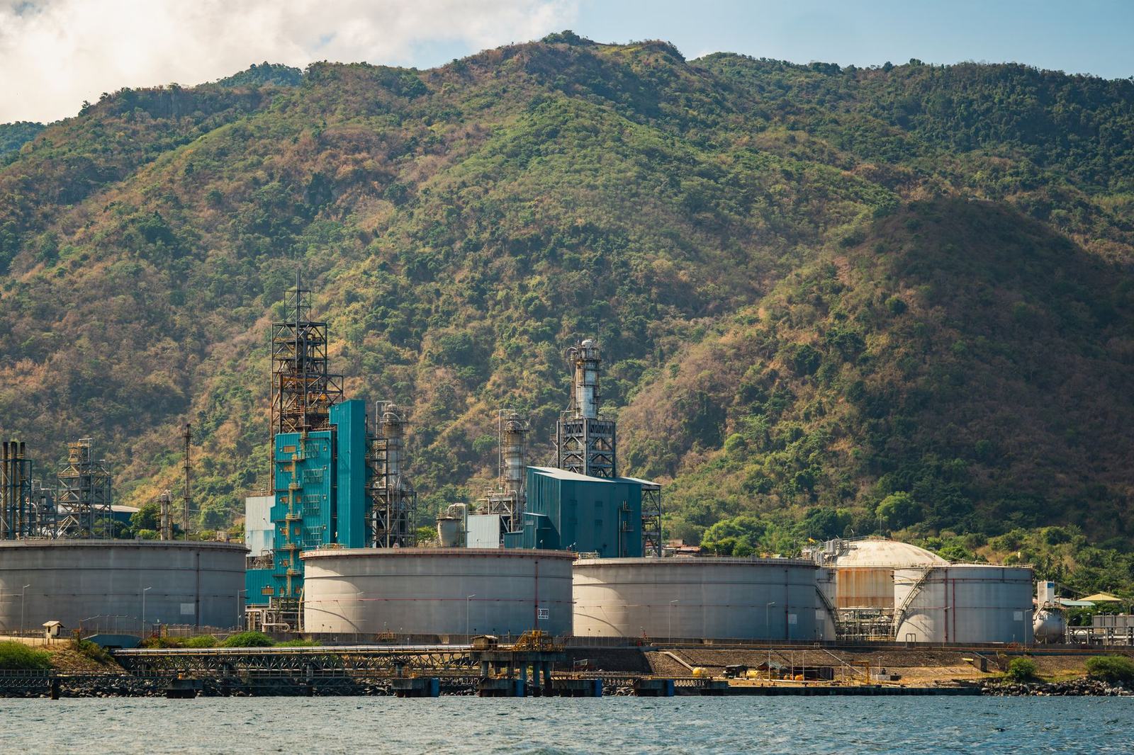 Coastal industrial plant with large storage tanks beneath green hills