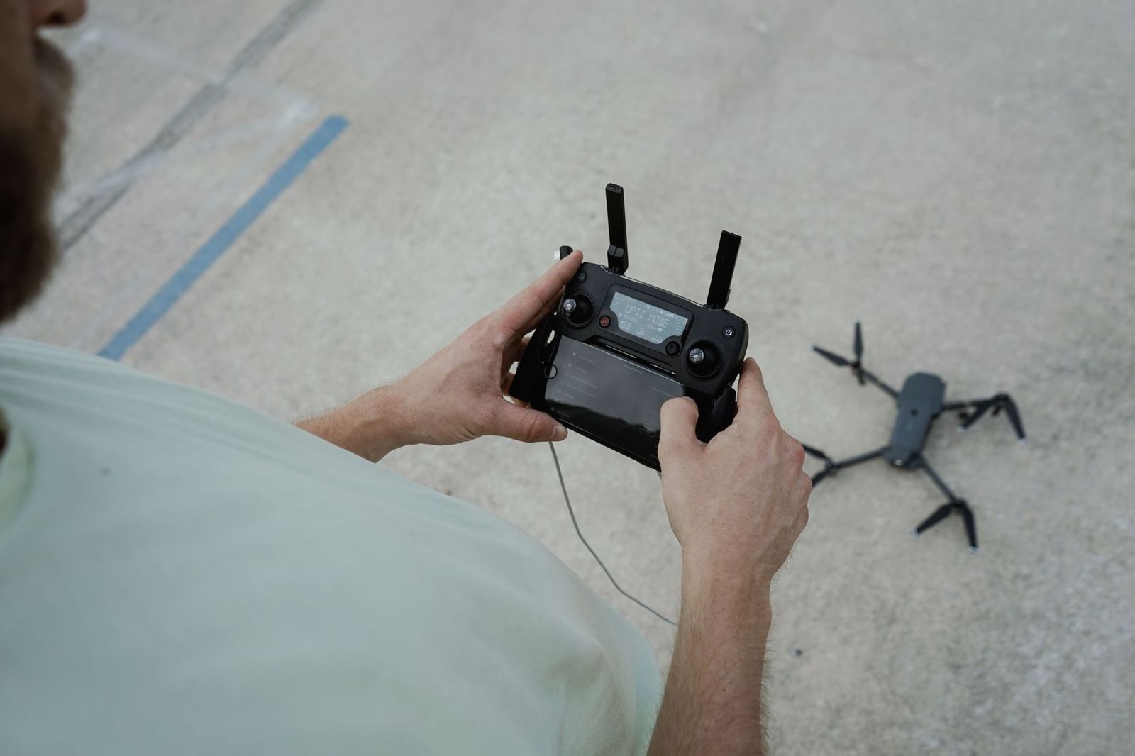 An operator holding a drone controller, with the drone resting on concrete beside him