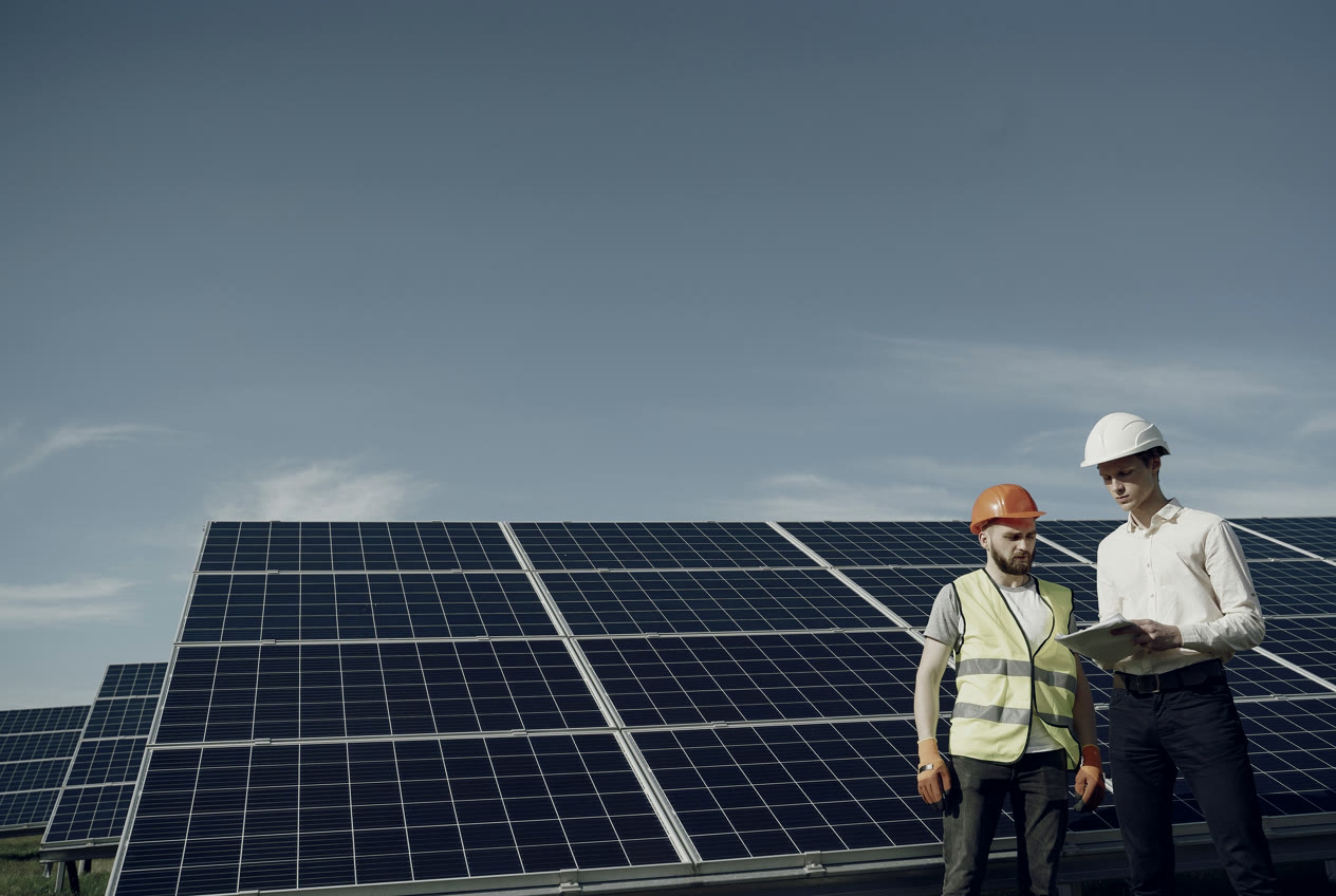 Two men in hard hats and a high-visibility vest reviewing plans in front of solar panels
