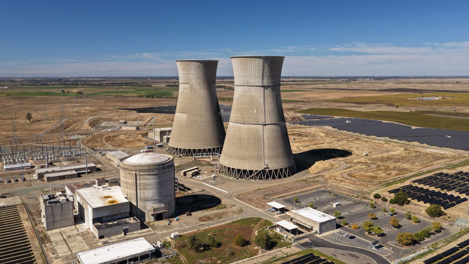 Aerial view of two power plant cooling towers with a substation and solar arrays around them
